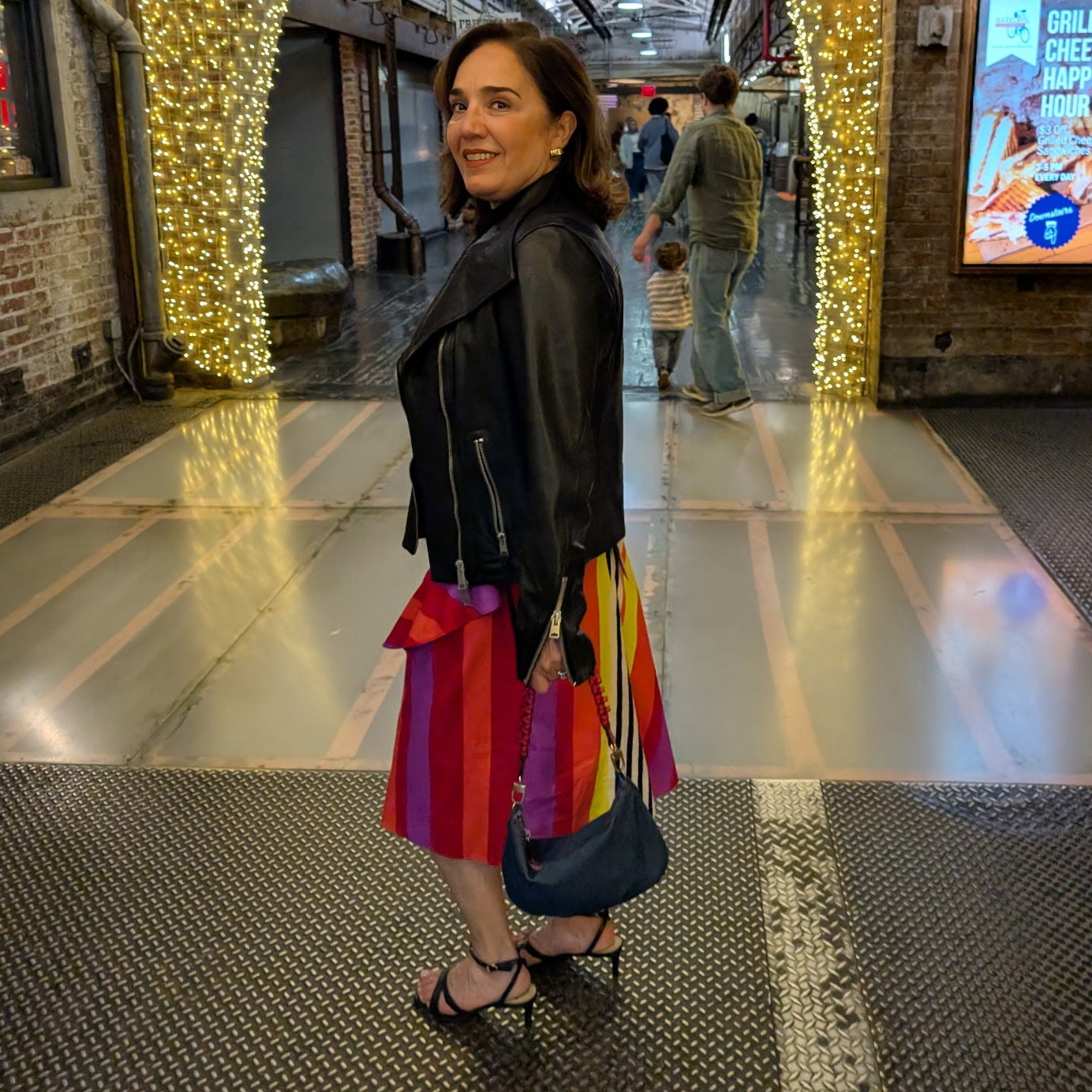 Woman in a colorful dress and black jacket standing in a modern indoor space with decorative lights.
